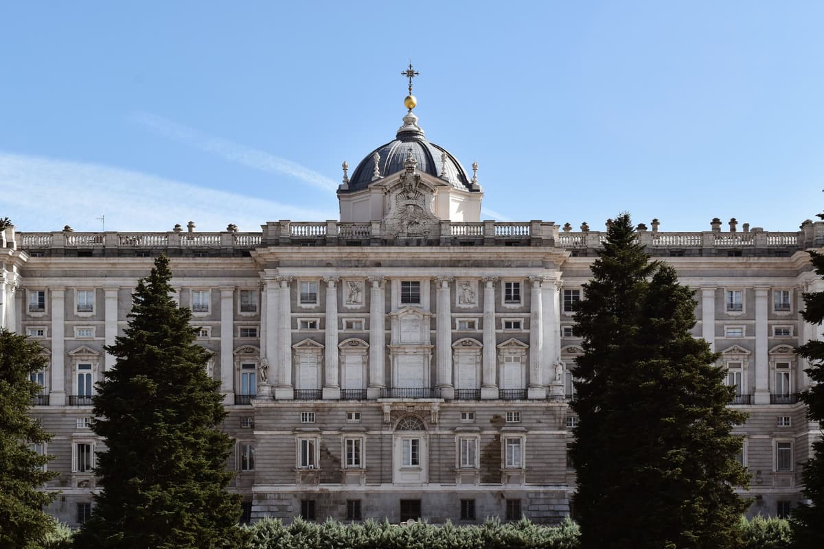 The Palacio Real de Madrid — the vast white limestone south facade above Plaza de la Armería, with the Almudena Cathedral domes to the right under a clear Castilian sky.